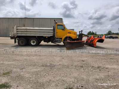 Dump truck with plow and orange snow piper on front, yellow Sterling LT8500 with cargo bed and trailer hitch in an open lot.