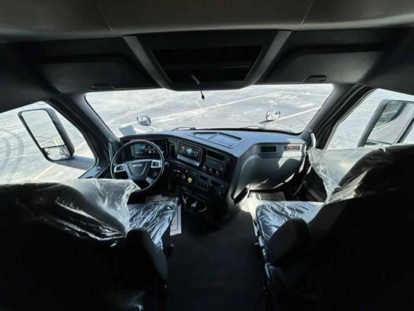 Cab interior of a 2023 Freightliner Cascadia 126 tractor, driver and passenger seats with plastic covers, dashboard with multiple gauges and screens, windshield with mirrors visible.