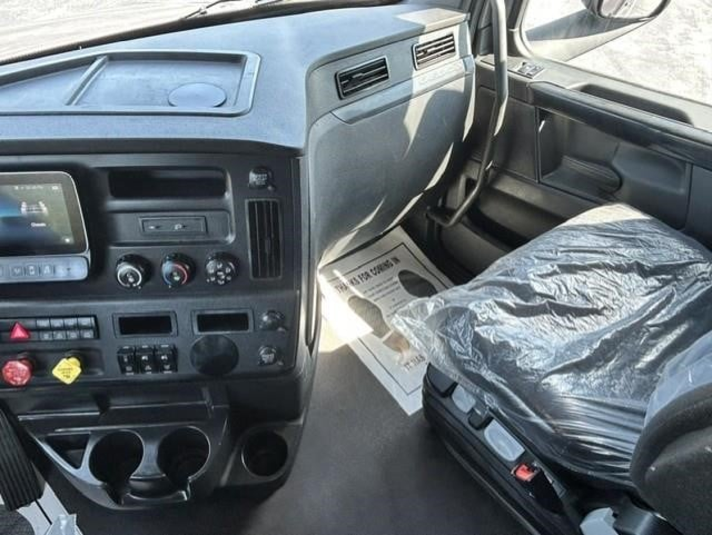 Cab interior of a 2023 Freightliner Cascadia 126 conventional sleeper showing steering wheel, dashboard controls, center console, and driver's seat with plastic cover.