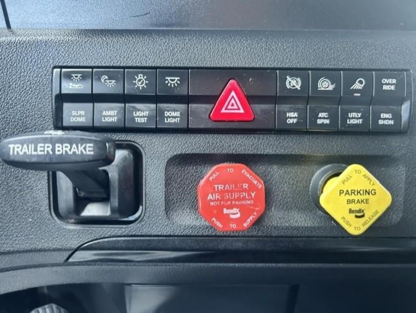 Freightliner Cascadia dash section showing hazard light switch and red air supply knob, yellow parking brake knob, trailer brake lever, and various indicator buttons.