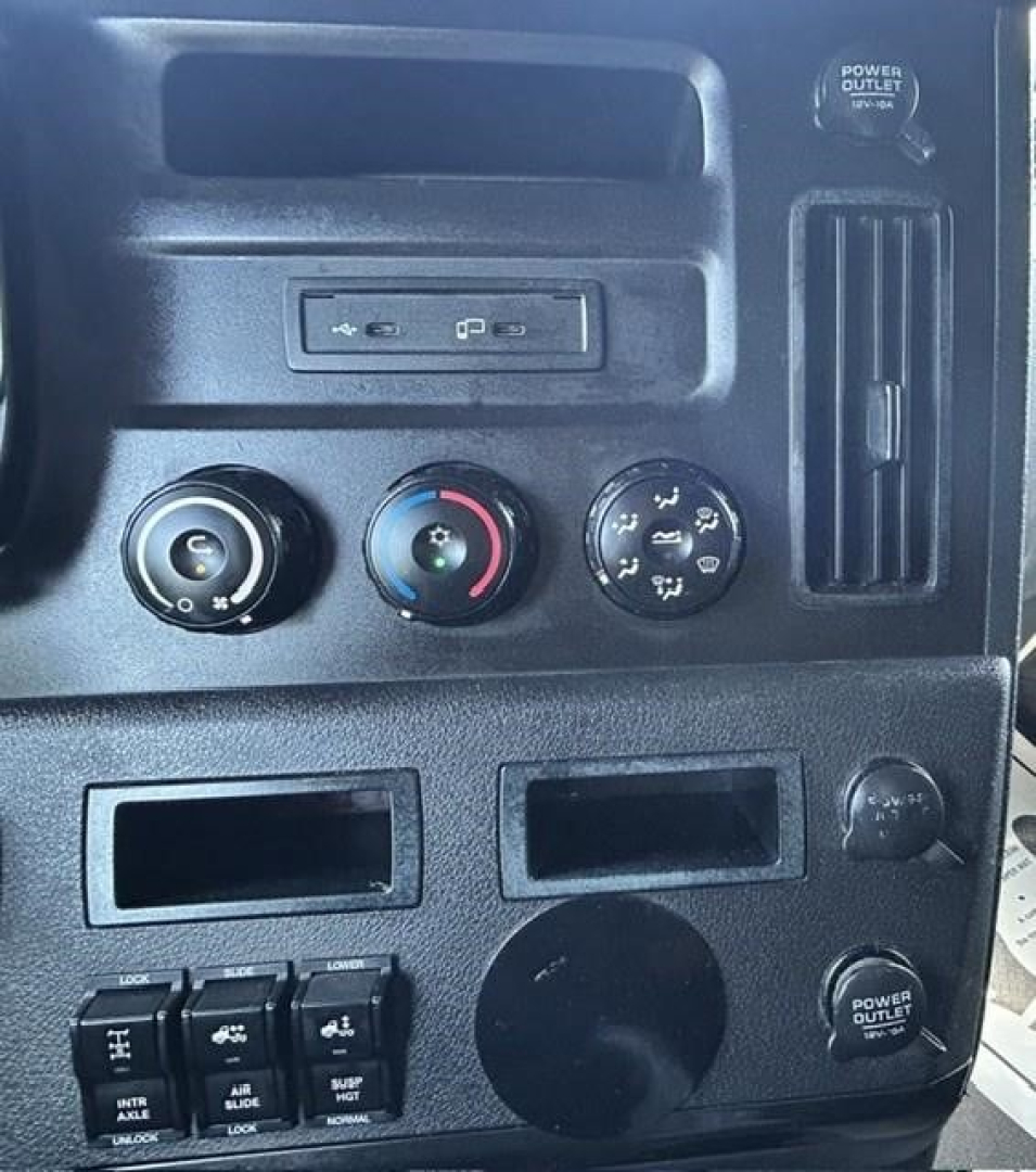 Dashboard of a Freightliner Cascadia showing climate controls, power outlets, and USB ports with various knobs and a circular blank panel.