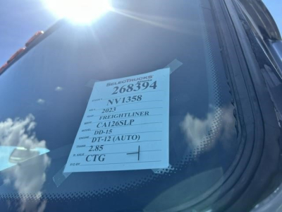 Truck window showing a white label with stock number 268394 and vehicle details: 2023 Freightliner Cascadia with model CA126LP, DD-15 engine, auto DT-12, 2.85 gear, VIN 1FUJHHDR4PLNV1358, CTG.