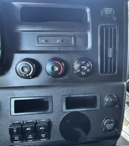 Dashboard of a Freightliner Cascadia showing climate controls, power outlets, and USB ports with various knobs and a circular blank panel.
