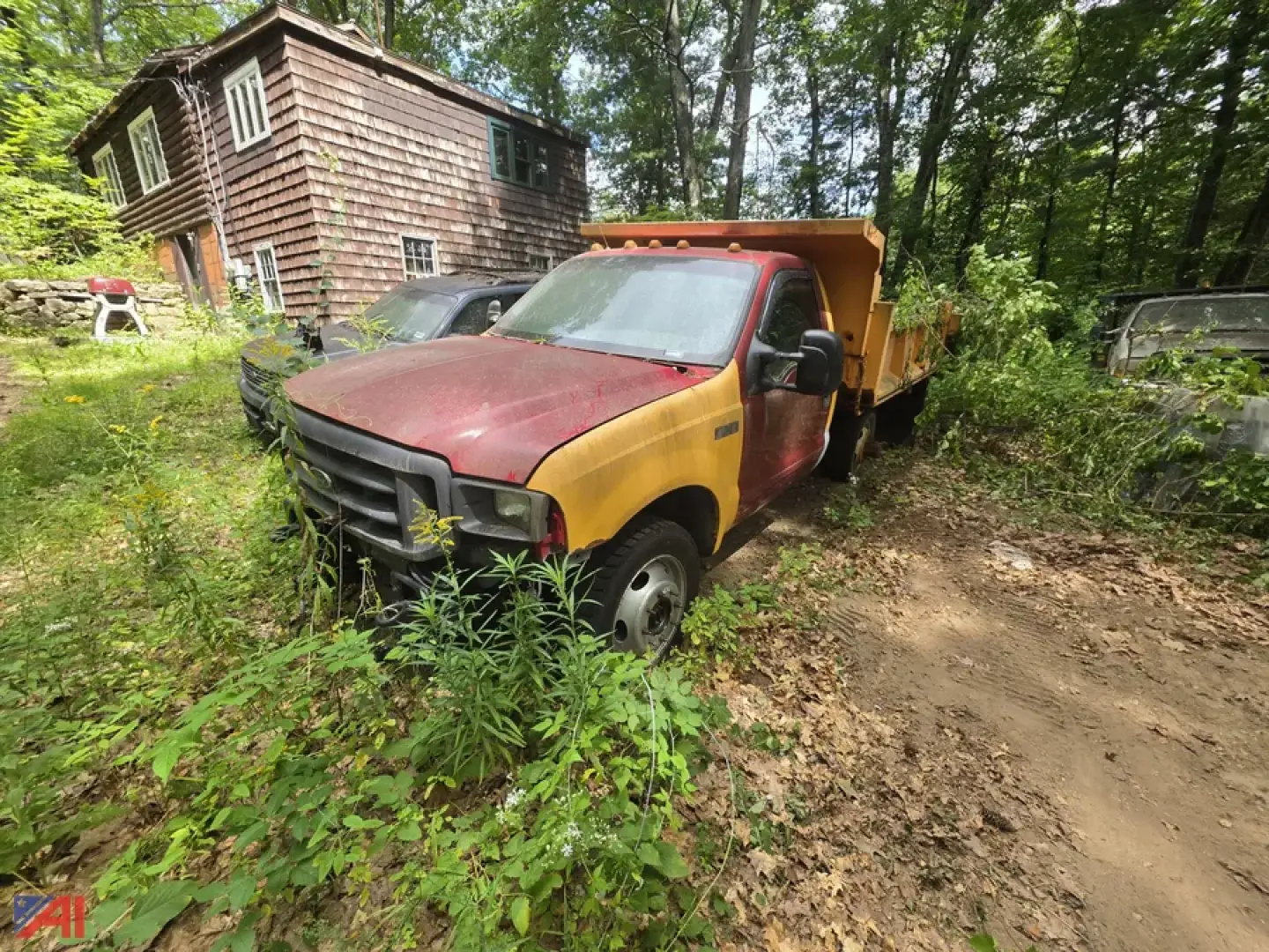 Dump truck in a wooded area showing a red cab and yellow dump bed, with overgrowth surrounding the vehicle and a wooden house in the background.