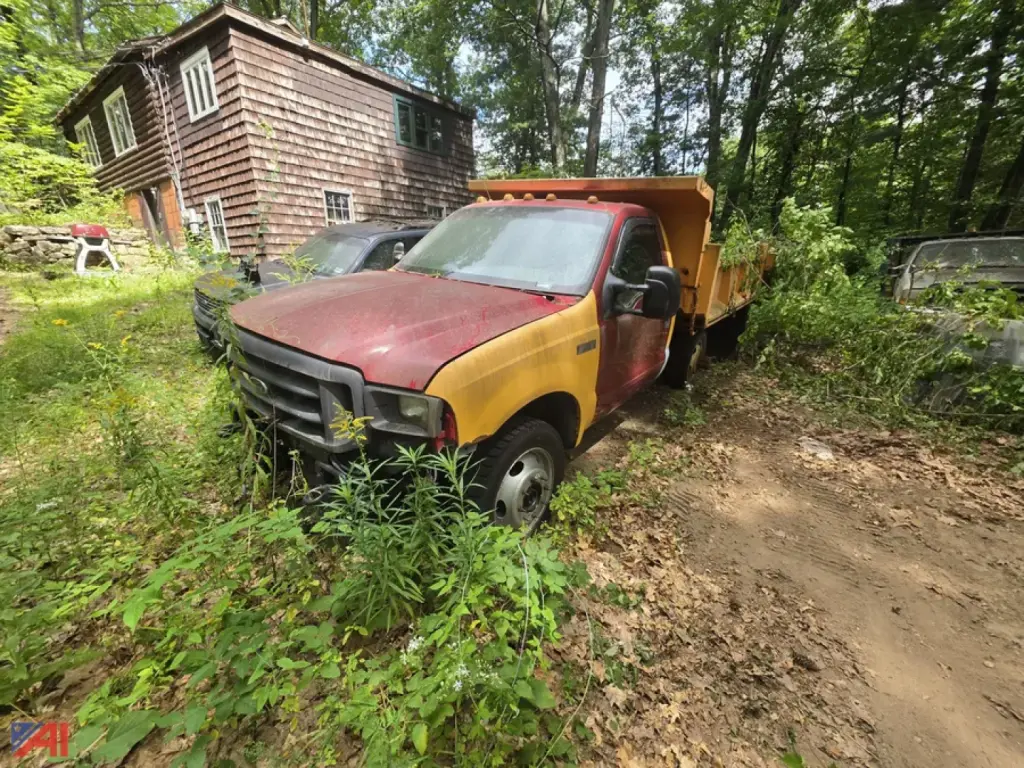 Dump truck in a wooded area showing a red cab and yellow dump bed, with overgrowth surrounding the vehicle and a wooden house in the background.