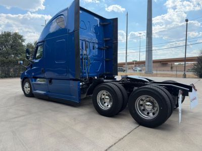 Truck cab and dual rear axles of a blue Freightliner semi-truck tractor in a parking lot, side view showing sleeper cab, rear wheels, and chassis.