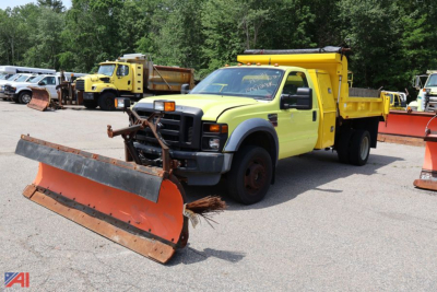 Ford F-450 Super Duty dump truck with front-mounted plow and yellow cab, snow-plow blade in foreground, other heavy equipment in background.