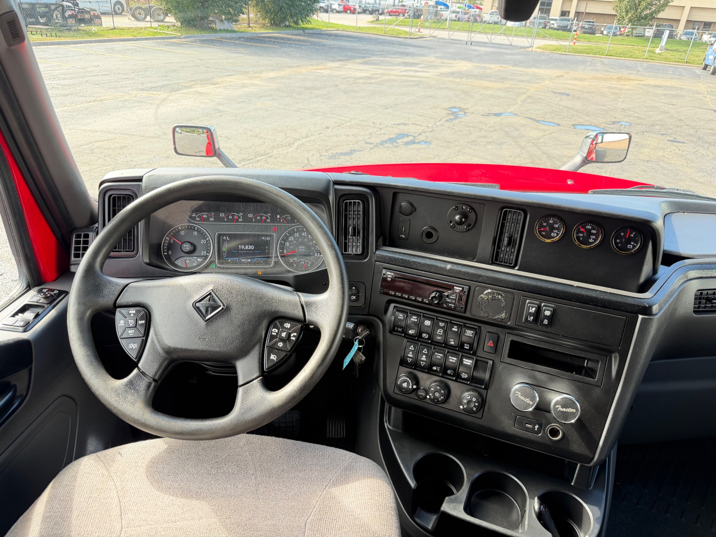 Vehicle cockpit showing steering wheel, dashboard, gauges, center console with controls, and red exterior/hood seen through windshield.