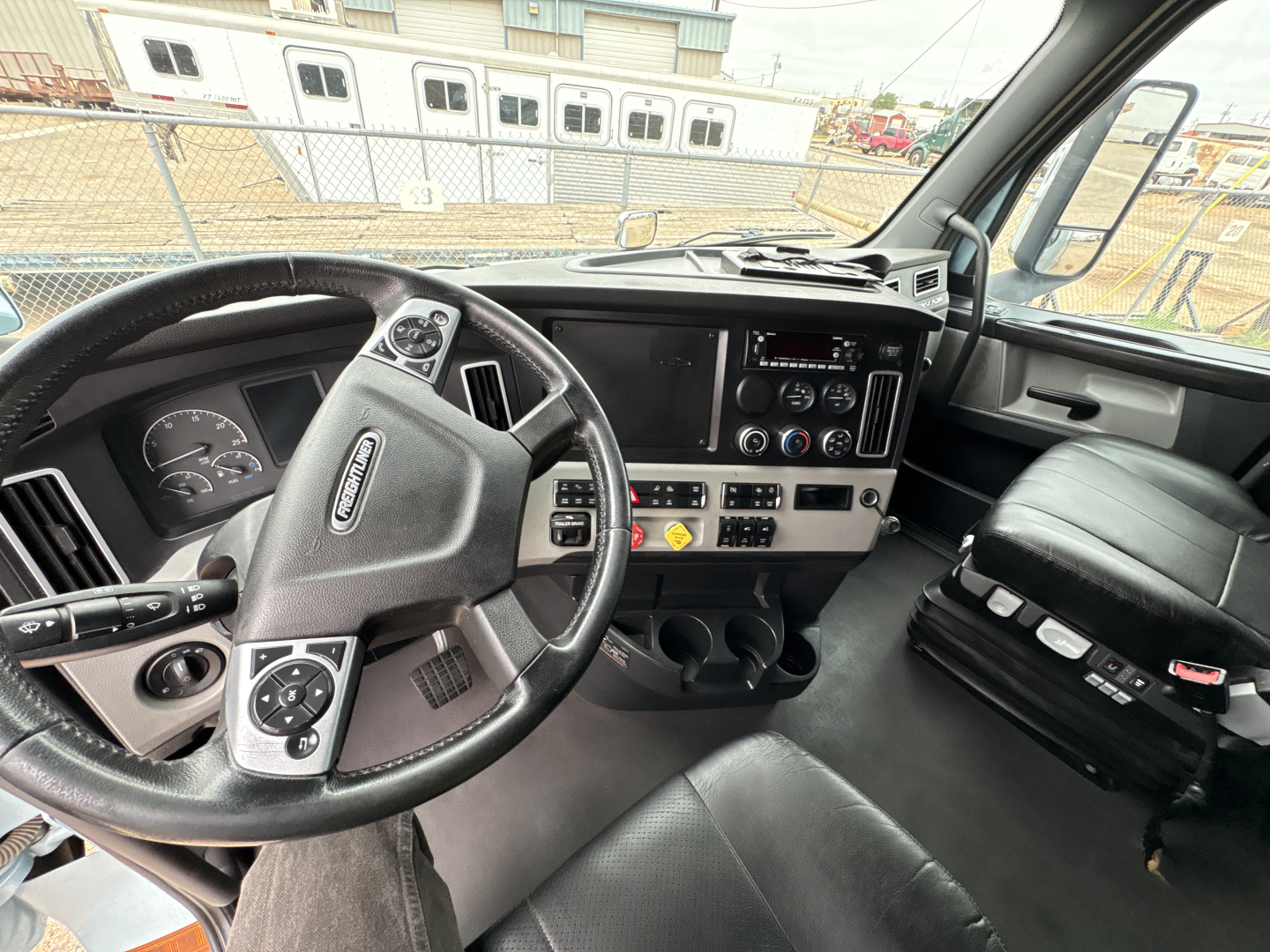 Truck cockpit showing steering wheel, dashboard, and center console of a 2023 Freightliner Cascadia 126. Visible gauges, controls, cup holders, and passenger seat.