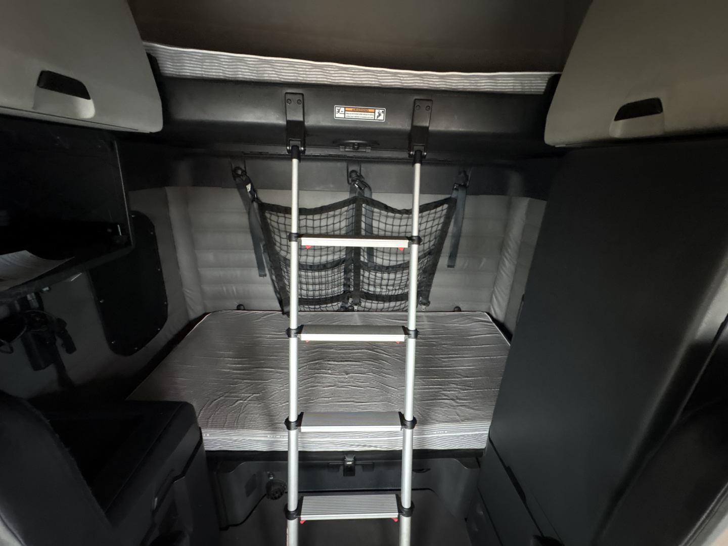 Freightliner Cascadia cab interior with fold-down bunk ladder, metal floor plate, and storage nets visible.