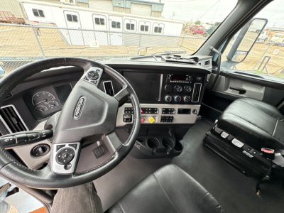 Truck cockpit showing steering wheel, dashboard, and center console of a 2023 Freightliner Cascadia 126. Visible gauges, controls, cup holders, and passenger seat.