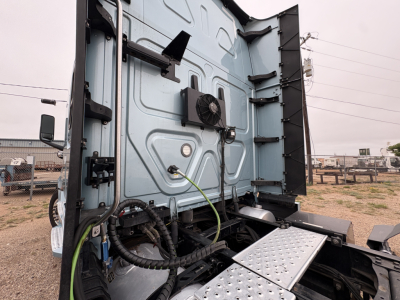 Image shows the rear cab area of a Freightliner Cascadia 126 truck with the rear doors open, exposing the cab interior and wiring loom. Exterior light blue body, with a hydraulic or electrical harness visible, and a diamond-plate step at th