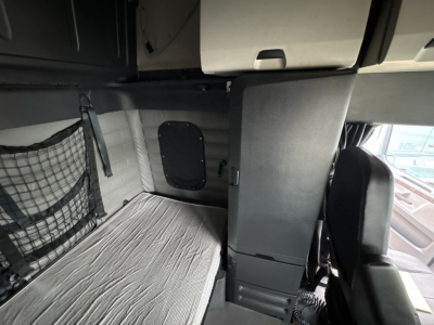 Cab interior of a Freightliner Cascadia 126 sleeper showing compact bunk with mattress and overhead storage cabinet. Passenger seat visible in foreground, side wall with ventilation panel, and a netted storage pocket.