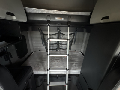 Freightliner Cascadia cab interior with fold-down bunk ladder, metal floor plate, and storage nets visible.
