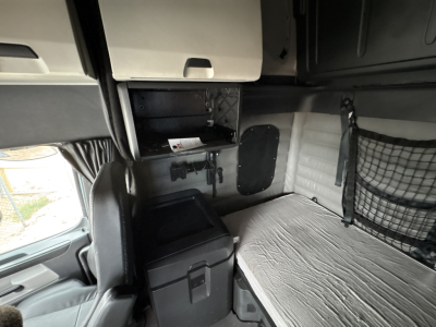 Cab interior of a 2023 Freightliner Cascadia 126 sleeper showing storage compartment, small fridge, and sleeping berth with netted pocket.