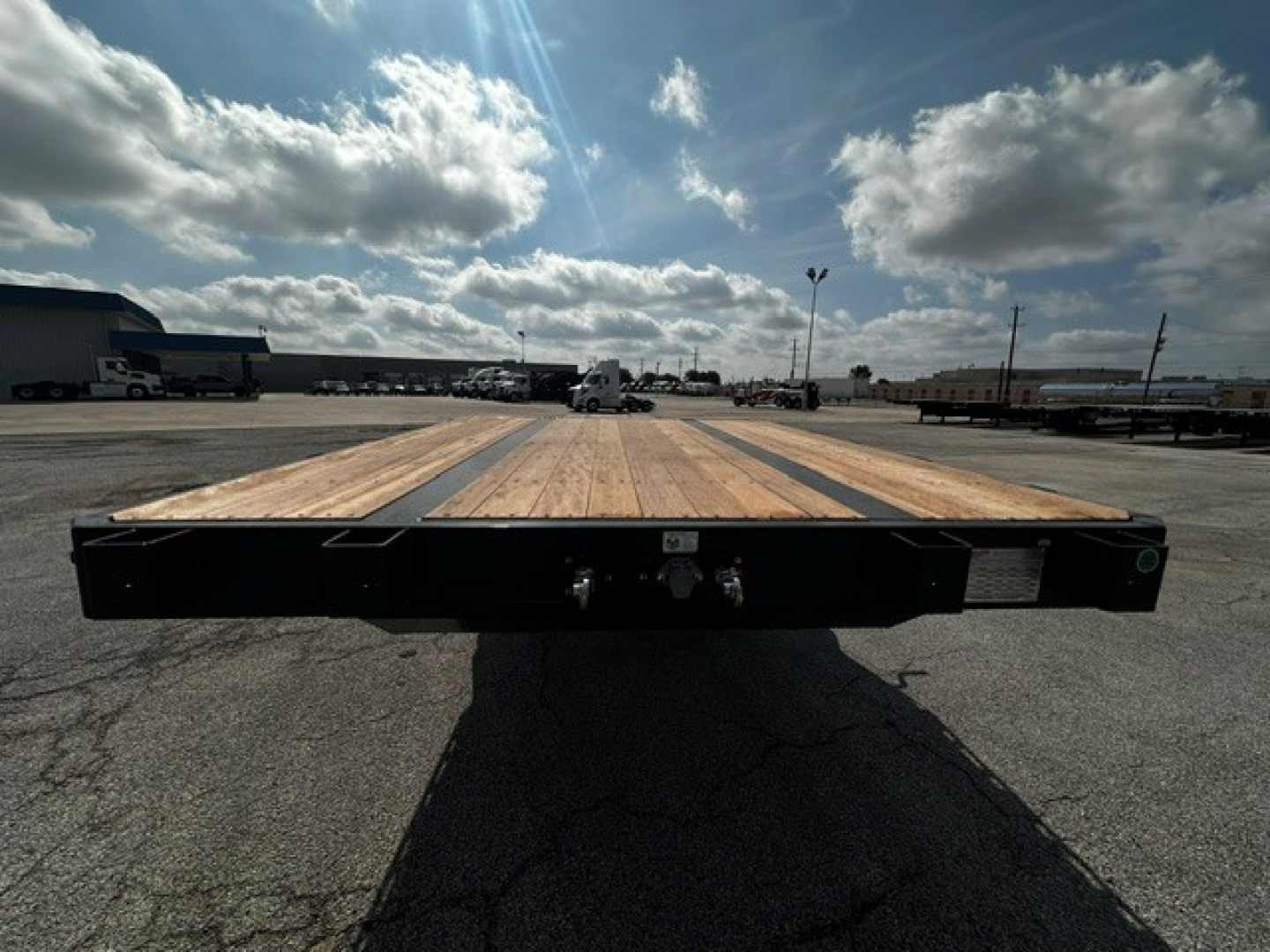 Flatbed trailer deck showing wooden deck planks and rear bumper with two latching ramps, black steel frame, clear sky in background.