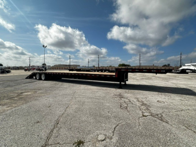 Long flatbed trailer with drop deck and closed tandem axles in an open lot, clear sky.