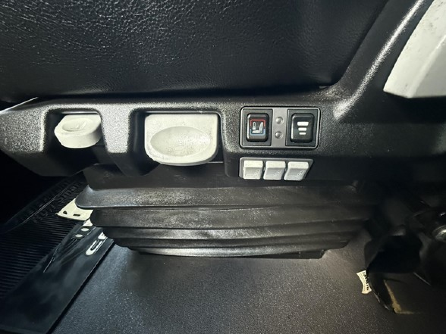 Cab interior under driver seat area showing control switches and knob with blank panel and four small buttons.