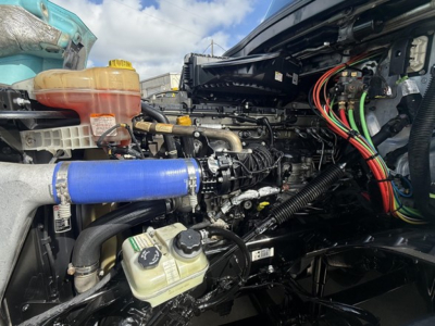 Truck engine bay of a 2023 Freightliner Cascadia 126 showing coolant reservoir, power steering/auxiliary lines, blue silicone intercooler hose, and various wiring harnesses.