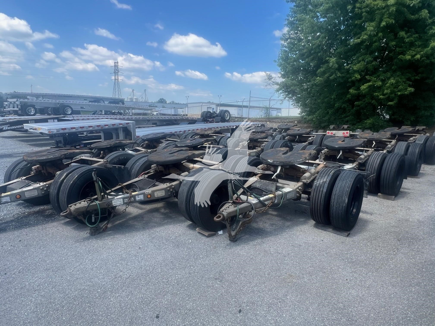Trailer dolly axles and wheel assemblies in a yard, multiple axles with tires and frames, blue sky and trees in background.