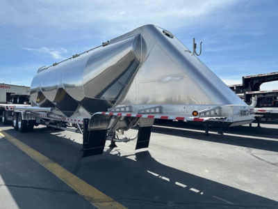 Pneumatic dry bulk trailer by Mac, shiny aluminum, side view showing cylindrical storage with rounded ends, tandem axles and rear discharge chute, equipment label details not legible.