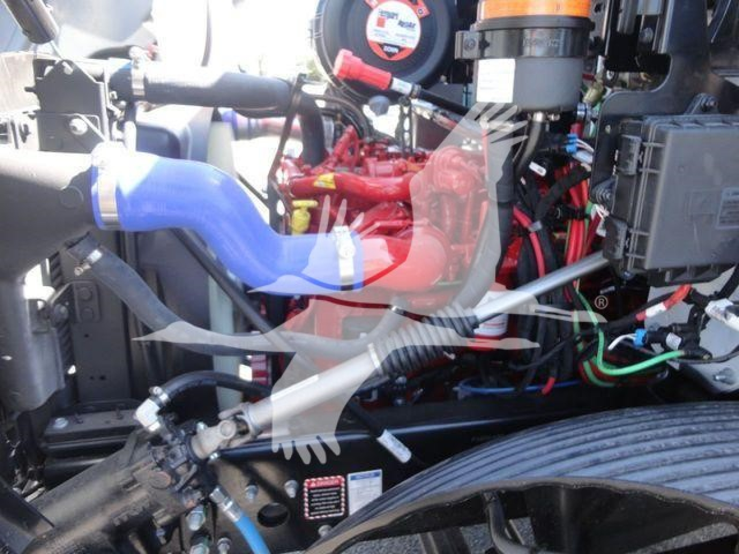 Truck engine bay of a 2027 Mack MD6 in an infrastructure bay showing red engine components and blue hoses with visible wiring and components.