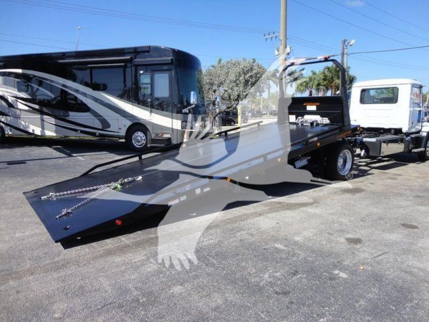 Truck bed with a low-angle view of a 2027 Mack MD6 rollback truck, flatbed with ramps, attached to a white tow vehicle in a dealership lot.