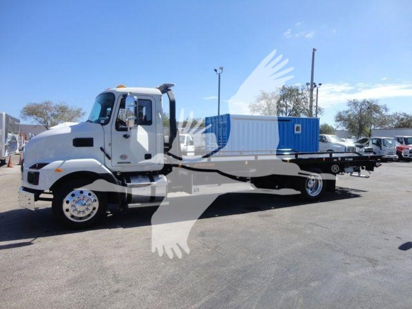Truck cab and flatbed rollback with blue container on a lot; clear sky.