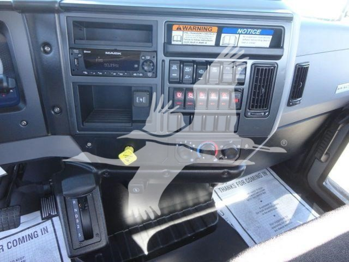 Dashboard area of a Mack MD6 truck showing steering wheel, radio, dashboard controls, warning labels, and pedals; central console with vents and climate controls visible.