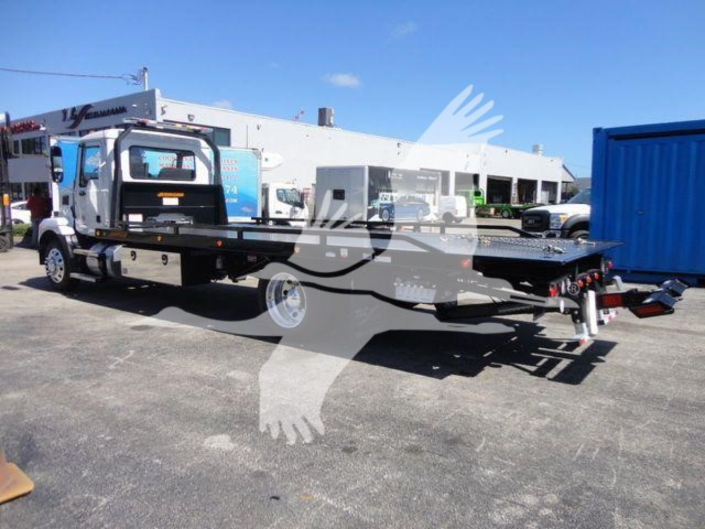 Flatbed rollback truck on lot with cab and long steel platform; Mack MD6 chassis visible with wheelbase, fuel tank, and rear ramps.
