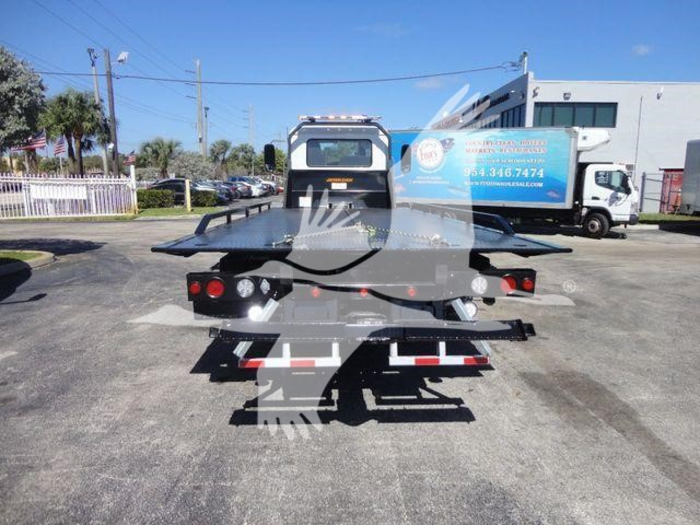 Tow truck in a dealership lot, rear view showing flatbed platform and red/white reflective markings.