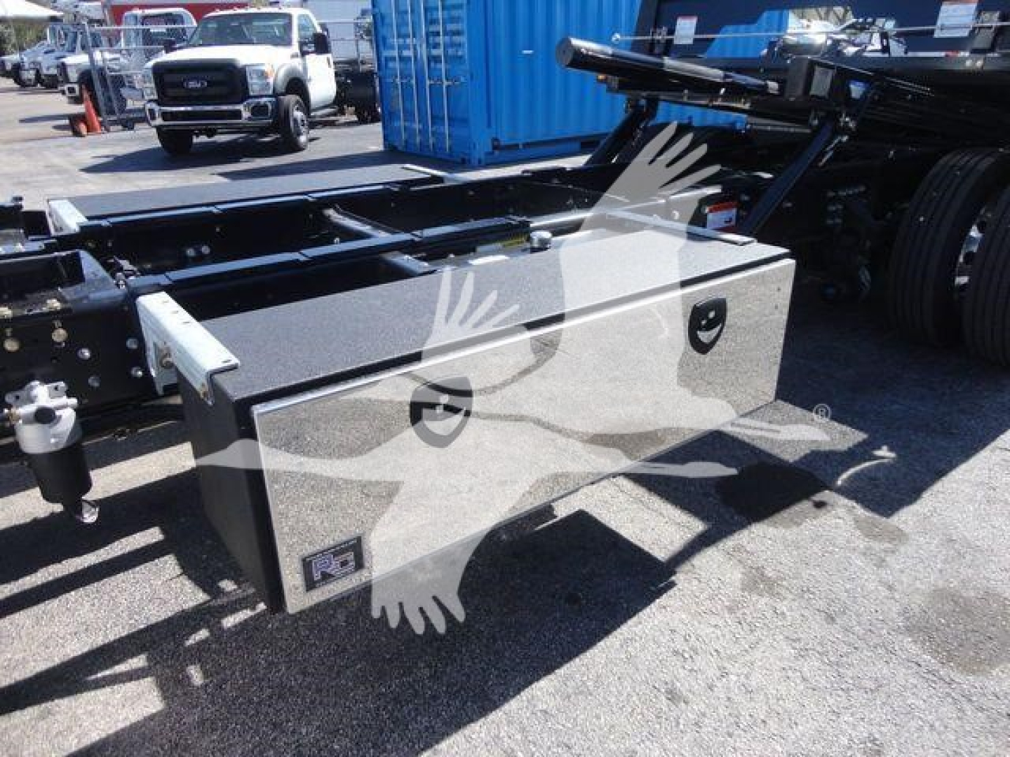 Two-compartment rectangular metal toolbox mounted near the frame of a Mack MD6 rollback truck. Visible text on toolbox not legible; background shows other trucks and equipment.