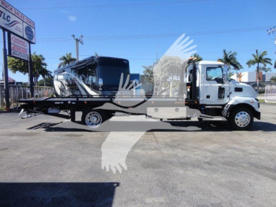 2027 Mack MD6 rollback truck with a white cab and a black flatbed, parked outdoors; showroom signage in background.