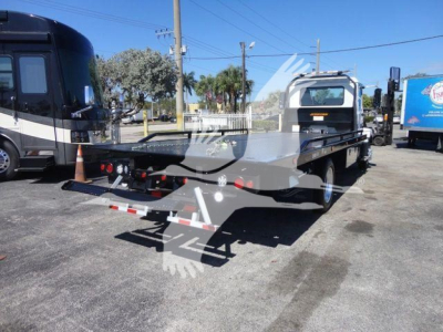 Flatbed rollback tow truck rear view showing hydraulic ramps and hitch, taillights, and yellow license plate area; Mack MD6 rolling chassis in a dealership lot.