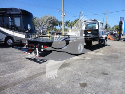 Flatbed tow truck on trailer with a white cab, loading ramp, and other trucks in background.
