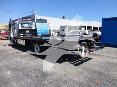 MD6 Mack rollback truck on a flatbed trailer with loading winch and wheel chocks in a dealership lot.
