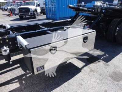 Two-compartment rectangular metal toolbox mounted near the frame of a Mack MD6 rollback truck. Visible text on toolbox not legible; background shows other trucks and equipment.