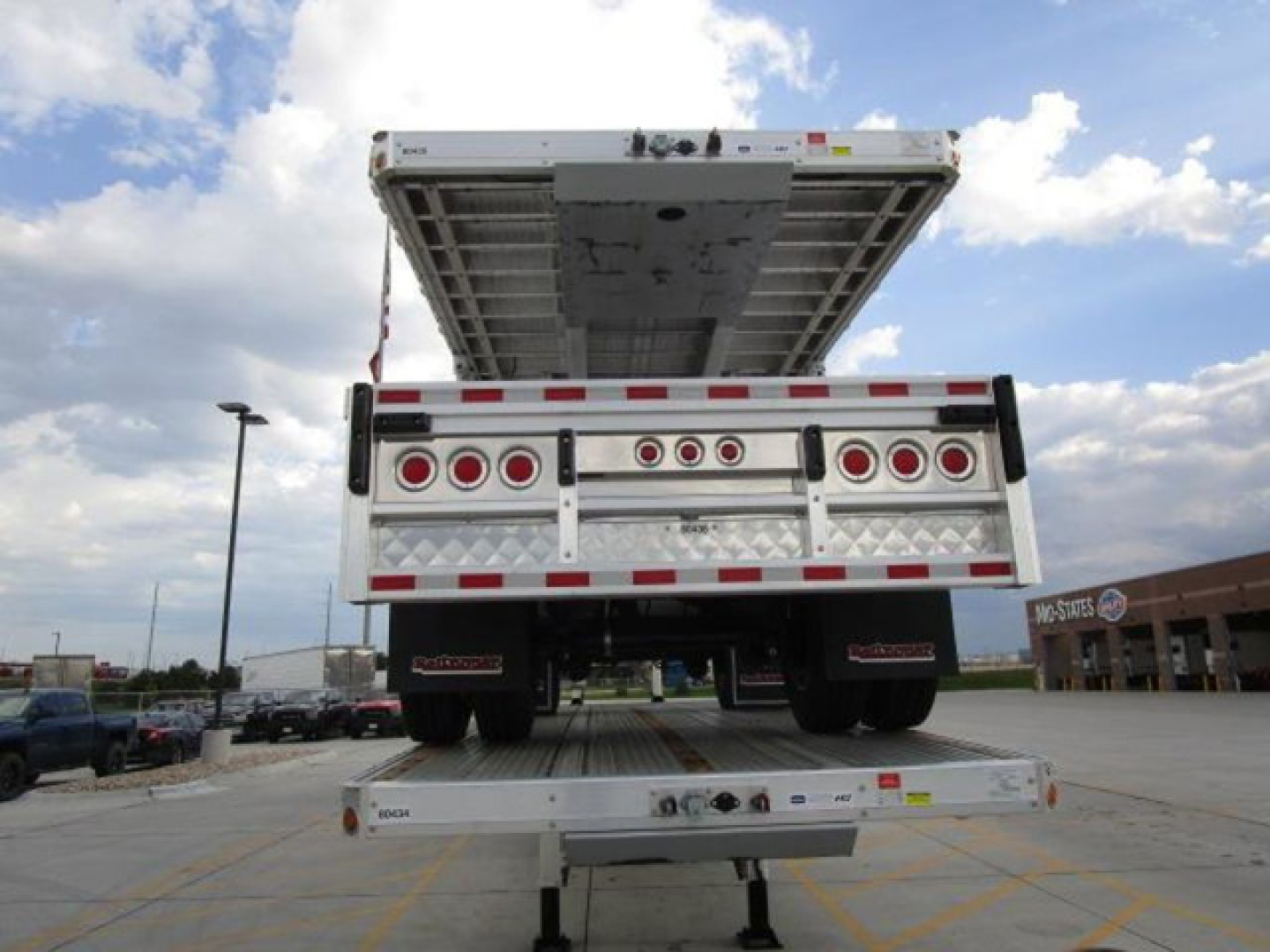 Rear view of a new 2027 Reitnouer flatbed trailer with its lift gate raised, showing taillights, reflective stripes, and loading deck.