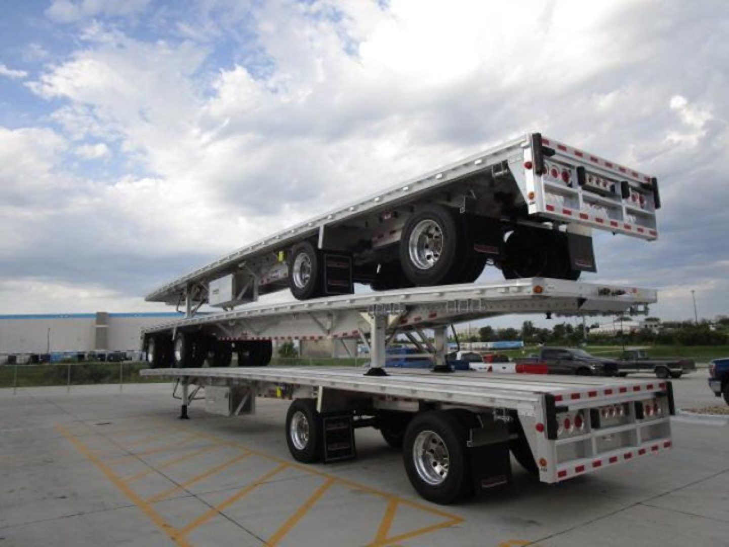 Flatbed trailer stacked on top of another at a dealership lot, with a spare tire and ramps visible; no legible branding or markings read clearly on the trailers.