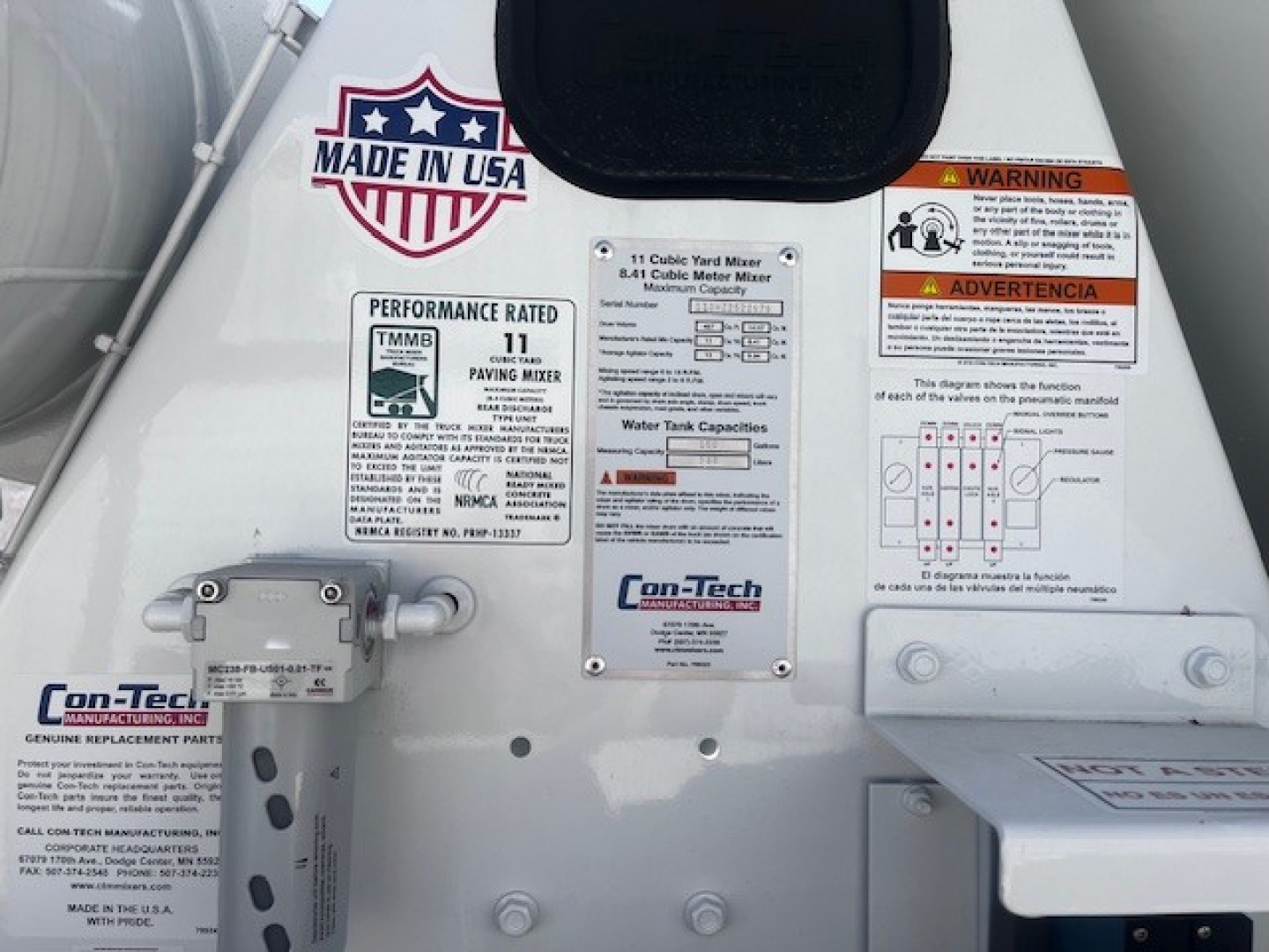 Mixer truck rear with labels: Made in USA badge, performance rating 11, Con-Tech/mixer manufacturer plate, warning and instruction diagrams, water tank capacity chart.