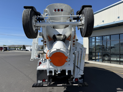 Front view of a white Mack GR84F mixer truck, rear wheels elevated, orange circular mixer housing, chassis and undercarriage components visible.