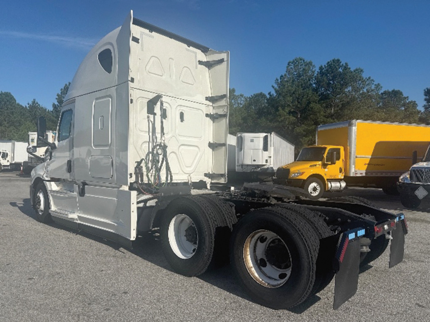 Truck tractor side/rear view showing white sleeper cab and dual rear axles with a yellow truck in background.
