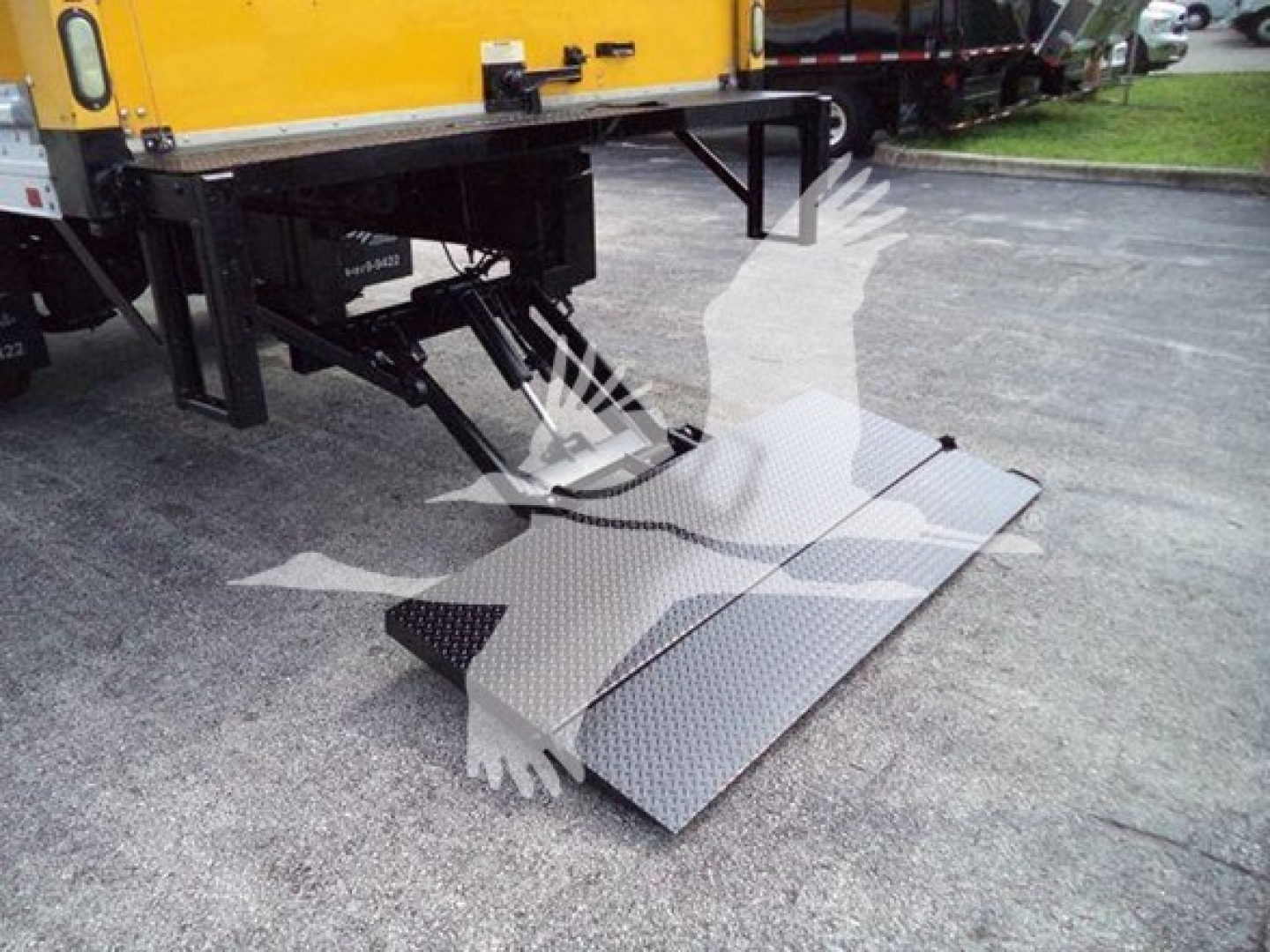 Truck side view with a yellow box truck and a fold-out aluminum ramp/platform at the rear for loading, showing metallic diamond-plate surface and hydraulic lift mechanism.