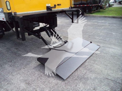 Truck side view with a yellow box truck and a fold-out aluminum ramp/platform at the rear for loading, showing metallic diamond-plate surface and hydraulic lift mechanism.