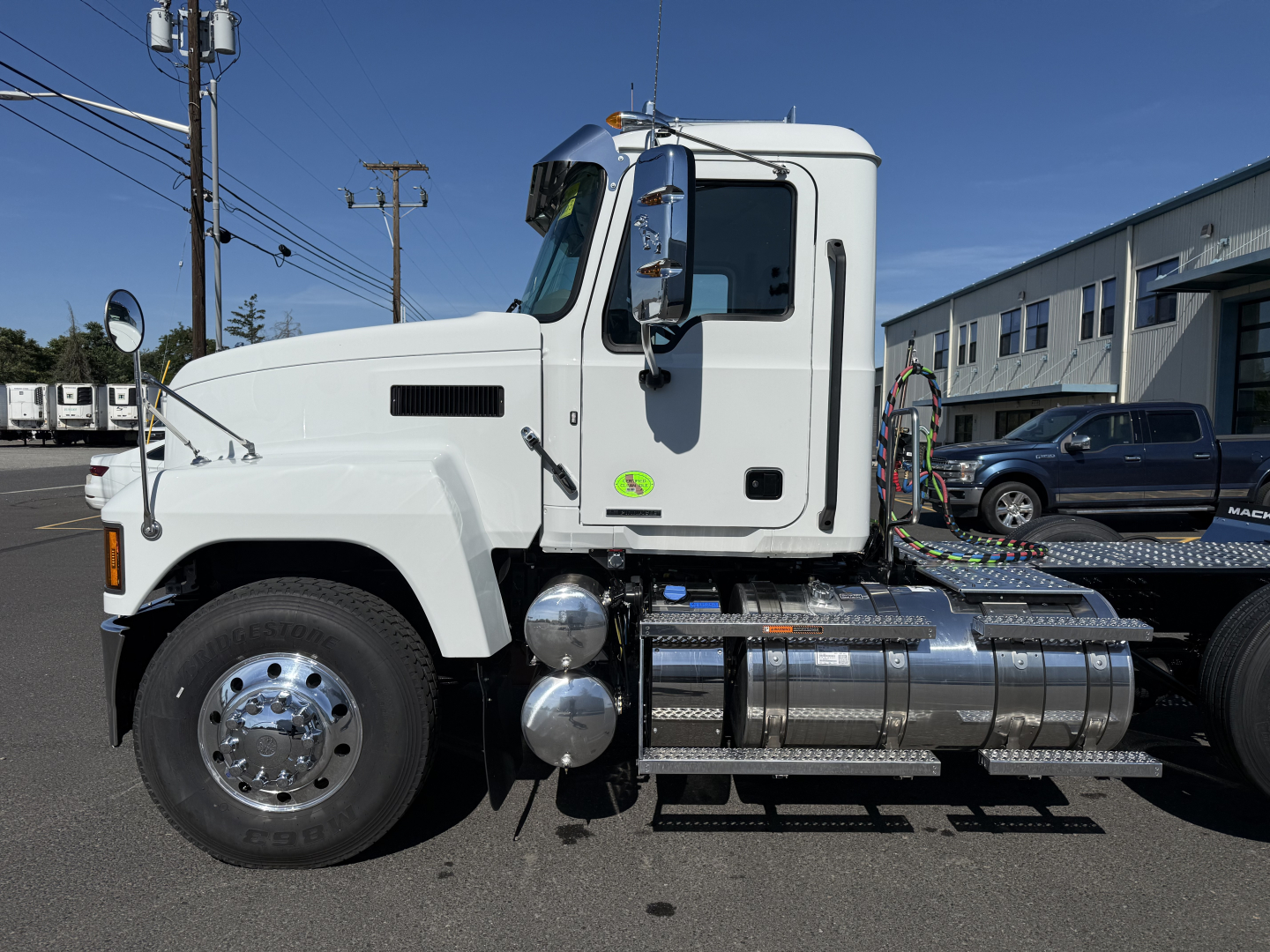 White 2027 Mack PI84T conventional daycab tractor side view showing chrome fuel tanks and steps, cab mirrors, front fender, and wheel; no text clearly visible on panels.