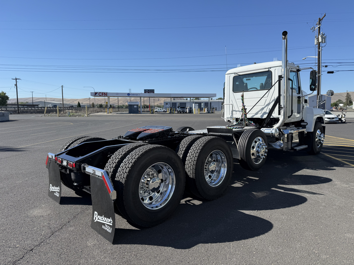 Tractor truck chassis and cab in a parking lot showing rear triple-axle configuration and rear mud flaps with brand text.