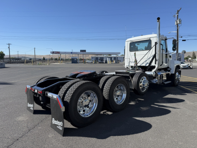 Tractor truck chassis and cab in a parking lot showing rear triple-axle configuration and rear mud flaps with brand text.