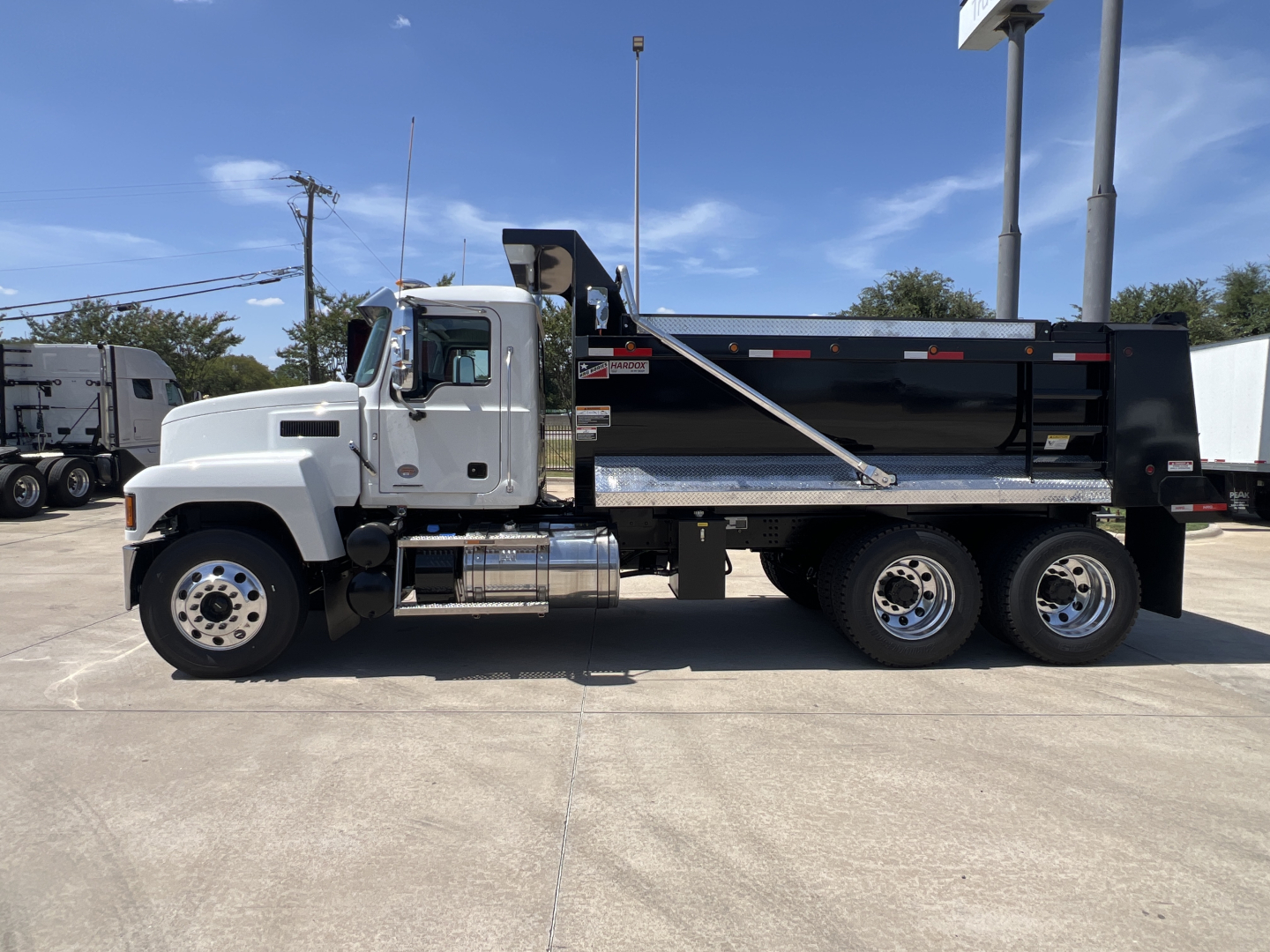 Dump truck side view showing 2027 Mack PI64 with a black dump bed and chrome fuel tanks; cab is white, three rear axles, diamond plate step, clear blue sky.