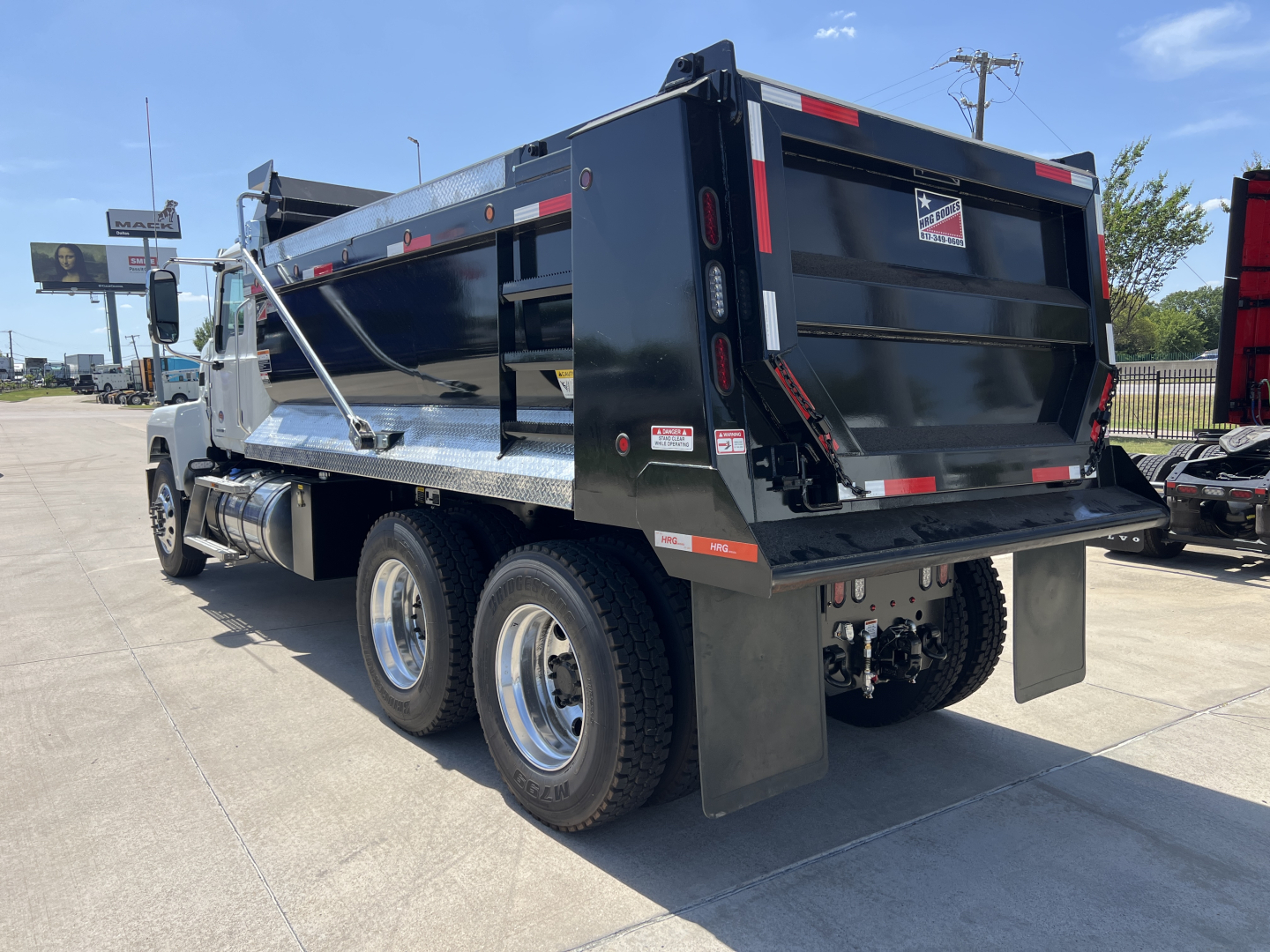 Mack PI64 dump truck rear left view showing dump bed and rear tires, with reflective safety stripes and bumper lights.