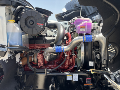 Engine bay of a 2027 Mack PI64 showing coolant reservoir, purple coolant overflow tank, radiator, turbocharger, hoses, and engine block with red paint.
